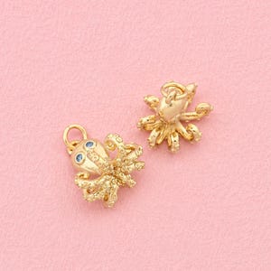 以下が含まれることがあります： 質感のある触手と目として小さな青い円形のアクセントが施された、ゴールドトーンのタコ型チャーム2点。各チャームの上部には取り付け用の小さな円形の丸カンが付いており、詳細な立体的な海の生き物のデザインが特徴です。