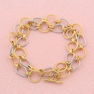 May include: A two-tone chain link bracelet featuring alternating gold-colored and silver-colored circular and oval links with a twisted rope texture. The bracelet is secured with a gold-colored toggle clasp consisting of a circular ring and a bar.