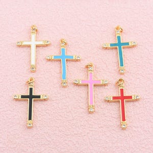May include: Six metal cross pendants featuring gold-tone frames and colorful enamel centers in white, light blue, dark blue, black, pink, and red. Each cross includes small clear rhinestones at the ends of the arms and a jump ring at the top.