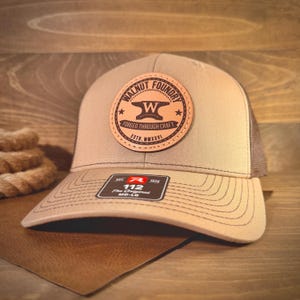 May include: A tan baseball cap with a brown mesh back and a circular leather patch on the front. The patch features the text WALNUT FOUNDRY, an anvil icon, and the text FORGED THROUGH CRAFT ESTD. MMXXVI. A black sticker on the brim reads 112.