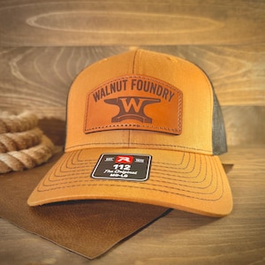 May include: A mustard yellow baseball cap featuring a brown leather patch on the front with the text WALNUT FOUNDRY and an anvil logo. The cap has a black mesh back and a black sticker on the brim with the text R 112 The Original MD-LG.