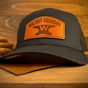 May include: A black trucker hat with a curved brim. The hat features a brown leather patch with the words "WALNUT FOUNDRY" and an anvil logo. The hat has a mesh back and is set against a wooden background.