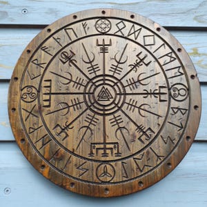 Puede incluir: Un adorno de pared redondo de madera con un acabado marrón envejecido. El diseño presenta intrincadas tallas de runas y símbolos nórdicos, incluyendo una brújula Vegvísir central. El borde exterior tiene pequeños agujeros redondos espaciados uniformemente.