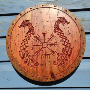 May include: A circular wooden shield with a Viking design. It features a central symbol flanked by two dragon-like figures. The wood has a reddish-brown hue with a darker brown border and is decorated with carved details.