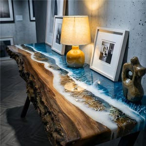 Peut inclure: Une console en bois faite à la main avec un bord brut et un motif de rivière en résine époxy bleue et blanche. La table présente une teinte de bois chaleureuse et est soutenue par des pieds noirs. Une lampe et des photos encadrées sont sur la table.