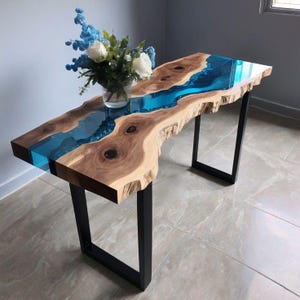 Pode incluir: Uma mesa de madeira com borda natural e um design de rio de resina azul. A mesa tem um grão de madeira natural e é suportada por pernas de metal preto. Um vaso de flores brancas e azuis está sobre a mesa.