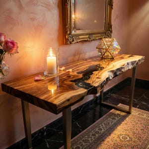 Könnte beinhalten: Eine Konsole aus Holz mit einer rohen Kante und einem dunklen Epoxidharzfluss. Der Tisch hat eine natürliche Holzmaserung und goldfarbene Metallbeine. Eine brennende Kerze in einem Glas und ein geometrisches Prisma befinden sich auf dem Tisch.