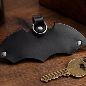May include: A black leather key organizer shaped like a bat silhouette with silver-toned metal rivets and a central circular ring attachment. A single brass-colored metal key is positioned separately on the surface next to the leather key holder.