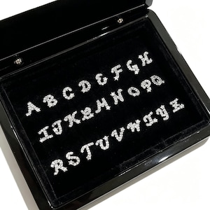 May include: A black jewellery box with a velvet interior displays a full alphabet of sparkling, silver-coloured letter charms. Each letter is encrusted with small, clear crystals, creating a dazzling effect. The box is open, revealing the neatly arranged letters.
