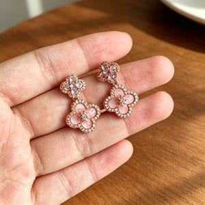May include: A pair of rose gold drop earrings featuring a clover-shaped design. Each earring consists of two stacked clover motifs inlaid with pale pink stone and bordered by small clear crystals, with a single crystal at the center of each.