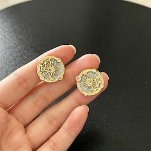 May include: A pair of gold-toned stud earrings. Each earring features a sunburst design with a central pink stone, surrounded by a white background and a gold border. Small clear and pink stones are set around the edge.