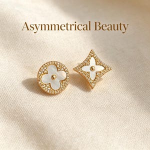 May include: Gold-toned asymmetrical earrings. One earring is a circle with a four-leaf clover design, the other is a diamond shape. Both have a white center and are encrusted with small, sparkling stones. The text "Asymmetrical Beauty" is above the earrings.