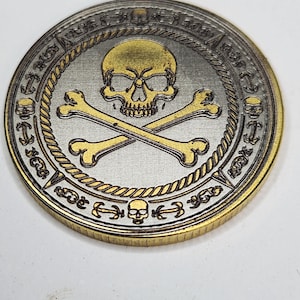 Aged metal coin with skull and crossed bones, shaded fills and gold rim, showing MOPA laser engraving effects.