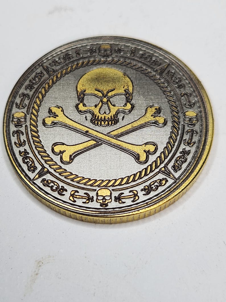 Aged metal coin with skull and crossed bones, shaded fills and gold rim, showing MOPA laser engraving effects.