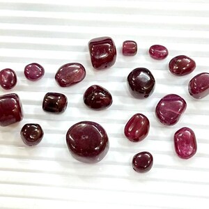 Puede incluir: Una colección de piedras preciosas pulidas, de color rojo intenso, de varios tamaños y formas, incluyendo formas redondeadas y rectangulares. Las piedras están dispuestas sobre una superficie blanca y estriada, mostrando su rico color y brillo.