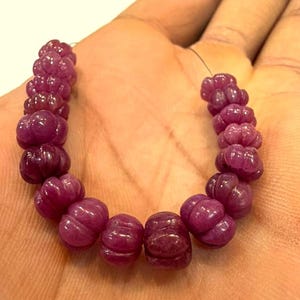 Può includere: Un filo di perline di pietre preziose magenta scuro scolpite. Ogni perla ha un design strutturato unico. Le perle sono infilate su un filo sottile e scuro. Le perle sono tenute nella mano di una persona.