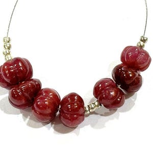 Può includere: Una collana di perline di pietre preziose scolpite, di colore rosso scuro. Le perline hanno la forma di zucca e sono infilate su un filo sottile color argento, con piccole perline distanziatrici argentate. Le perline hanno una finitura lucida.