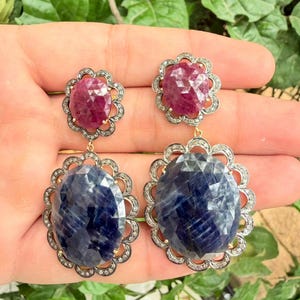 Op de afbeelding: Een paar oorbellen met gefacetteerde ovale blauwe saffieren en kleinere gefacetteerde ovale robijnen. Elke edelsteen is gezet in een geschulpte rand bezet met kleine witte diamanten, wat zorgt voor een gelaagd ontwerp met een bloemachtig uiterlijk.
