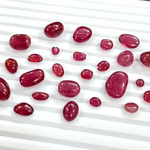 Puede incluir: Una colección de piedras preciosas pulidas, de color rojo intenso, de varios tamaños y formas. Las piedras están dispersas sobre una superficie blanca y acanalada, mostrando sus superficies lisas y reflectantes. Las gemas varían desde pequeñas cuentas redondas hasta piedras más grandes, de forma ovalada.