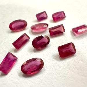 Könnte beinhalten: Eine Sammlung von zehn losen, facettierten Edelsteinen in tiefen Magenta- und Rubinrottönen. Die Steine haben verschiedene Formen, darunter ovale, rechteckige und quadratische Schliffe, die alle eine polierte, reflektierende Oberfläche aufweisen.
