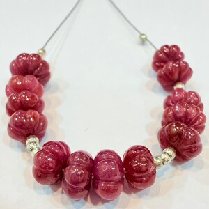May include: A strand of carved ruby beads. The beads are a deep red color and have a floral or pumpkin-like shape. The beads are strung on a thin, silver-colored wire.