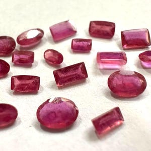 Könnte beinhalten: Eine Sammlung loser, facettierter Edelsteine in verschiedenen Schattierungen von tiefem Pink und Rot. Das Sortiment umfasst mehrere Formen wie ovale, rechteckige und quadratische Schliffe, die alle eine durchscheinende Qualität und polierte Oberflächen aufweisen.
