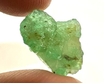 Könnte beinhalten: Ein roher, durchscheinender Smaragd-Edelstein mit leuchtend grüner Farbe. Das rohe Mineral weist eine kristalline Struktur auf und wird zwischen zwei Fingern vor weißem Hintergrund gehalten. Der Edelstein ist etwa 2,5 cm lang.