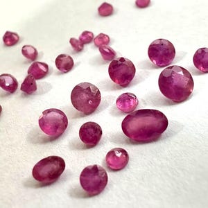 Op de afbeelding: Een verzameling losse, gefacetteerde edelstenen in verschillende tinten levendig roze en dieprood. Deze kleine, gepolijste stenen hebben ronde en ovale vormen met meerdere reflecterende oppervlakken die het licht opvangen en verspreid liggen.
