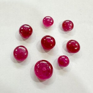 May include: A collection of eight polished, round, deep red gemstones. The gems vary slightly in size and are arranged on a white surface. The stones have a glossy, reflective surface, suggesting they are rubies or similar precious stones.
