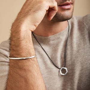 May include: A silver bracelet and a geometric pendant necklace. The bracelet is a simple, thin band. The necklace features a silver, six-sided pendant on a black cord. The person is wearing a light brown sweater.