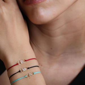 Puede incluir: Tres pulseras personalizadas con cierres dorados y dijes con iniciales. Las pulseras están ensartadas en cordones rojos, negros y turquesa. Los dijes con iniciales son las letras "E", "M" y "A".