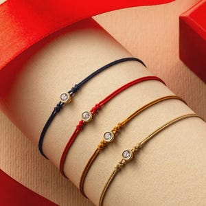 May include: Four delicate bracelets with a single, sparkling gemstone set in gold. The bracelets feature navy, red, mustard, and tan cords. A red ribbon and a red box are in the background.