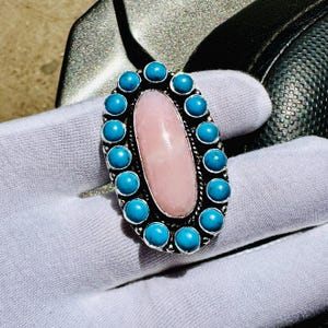 May include: Oval-shaped silver ring featuring a large, light pink center stone. The ring is encircled by a row of turquoise-colored, round stones. The ring is set on a white surface.