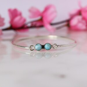 May include: A delicate silver bracelet featuring two round, light blue gemstones. The bracelet is thin and elegant, with a simple design. Pink flowers are blurred in the background, adding a soft, feminine touch to the image.