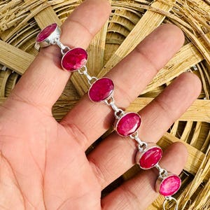 Puede incluir: Una pulsera de plata con piedras preciosas rojas vibrantes de corte ovalado. La pulsera está compuesta por engastes individuales de piedras preciosas conectados por pequeños eslabones de plata. Las piedras son de un color rojo intenso.