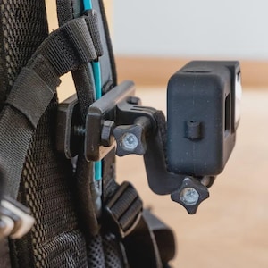 May include: A black action camera mounted on a backpack strap. The camera is attached with a black bracket and star-shaped screws. The backpack is black with a mesh design and black straps.