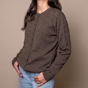 Könnte beinhalten: Ein brauner Cardigan mit Knöpfen und langen Ärmeln. Der Cardigan ist aus einem strukturierten Stoff gefertigt und hat einen runden Ausschnitt. Die Knöpfe sind klein und passen zur Farbe des Cardigans. Die Person trägt eine blaue Jeans.