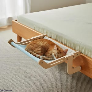 Op de afbeelding: Een slapende oranje kat ligt op een lichtblauw en wit kattenbed dat aan een houten bedframe is bevestigd. Het kattenbed heeft een hangmat-achtig ontwerp, met lichtblauwe stof gespannen tussen houten steunen. Het bed bevindt zich in de buurt van het voeteneinde van het bed.