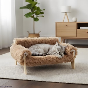 May include: A small, brown, fluffy pet sofa with wooden legs. A grey and white cat is sleeping peacefully on the bed. The bed is on a light-coloured rug, with a wooden cabinet and plant in the background.