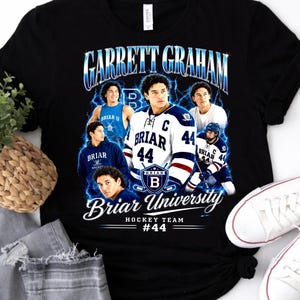 Può includere: Una t-shirt nera a maniche corte con una stampa grafica di Garrett Graham e della squadra di hockey della Briar University. Il design include diverse immagini di un giocatore di hockey, accenti di fulmini blu e il testo "Garrett Graham Briar University Hockey Team #44".