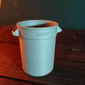 以下が含まれることがあります： 側面に2つのハンドルがあり、前面に「BRUTE」という文字が浮き彫りになった、円筒形の小さなライトグレーのプラスチック製ゴミ箱。容器は滑らかなマット仕上げで、上部が大きく開いており、標準的なゴミ箱のデザインを模しています。