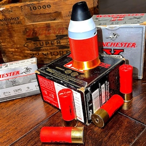 May include: A red and white plastic object resembling a shotgun shell sits atop a black Winchester ammunition box. Several red shotgun shells with brass bases are arranged around the box, alongside two additional Winchester boxes and a wooden crate.