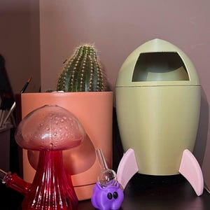 May include: A red mushroom-shaped glass water pipe and a purple dab rig with a clear glass top and black eyes. A light green rocket-shaped container with pink fins sits next to a cactus in a peach-colored pot.