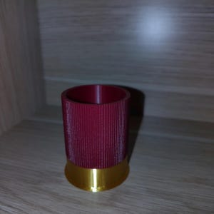 May include: A cylindrical container designed to resemble a shotgun shell, featuring a deep red, vertically ribbed upper section and a smooth, metallic gold-coloured base. The object has an open top and a flat bottom, standing upright as a single unit.