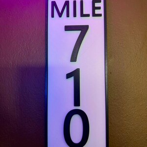 May include: A rectangular sign with the word "MILE" at the top, and the numbers "710" below. The text is black and raised against a white background, framed by a black border. The sign is likely for road or trail marking.