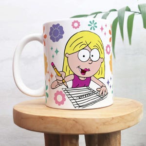 May include: White ceramic mug with a cartoon illustration of a blonde girl writing. The mug is decorated with colourful flowers and diamonds. The girl wears a pink top and holds a pencil. The mug has a white handle.