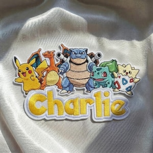 Può includere: Una toppa ricamata che riporta il nome Charlie in lettere gialle con un bordo bianco. Sopra il nome ci sono cinque colorati personaggi dei cartoni animati: un Pikachu giallo, un Charizard arancione, un Blastoise blu, un Bulbasaur verde e un Togepi.