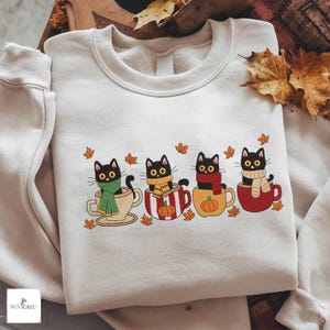 Op de afbeelding: Een crèmekleurige sweater met ronde hals en een grafische print van vier zwarte katten die in kleurrijke mokken zitten. Elke kat draagt een sjaal in een andere kleur en het ontwerp bevat kleine oranje herfstbladeren rondom de mokken.