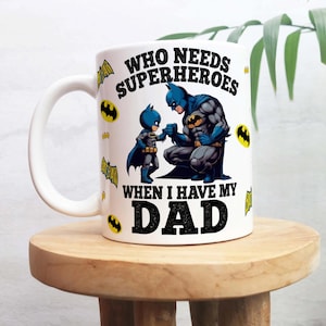 Puede incluir: Taza de cerámica blanca con un diseño de Batman. La taza muestra a Batman y a un niño con un disfraz de Batman, con el texto "WHO NEEDS SUPERHEROES WHEN I HAVE MY DAD". La taza tiene un asa blanca y está decorada con logotipos de Batman.
