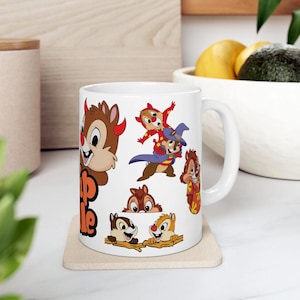 Può includere: Tazza in ceramica bianca con immagini dei cartoni animati di Cip e Ciop. La tazza ha un manico grande ed è decorata con varie immagini dei personaggi in diverse pose. La tazza è appoggiata su un sottobicchiere quadrato.